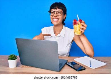 Beautiful brunettte woman working at the office eating healthy fruit pointing finger to one self smiling happy and proud 