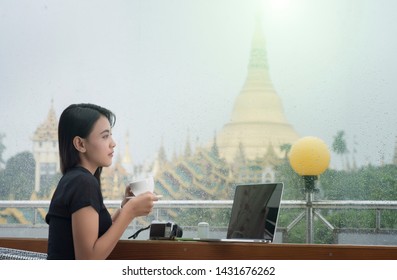 Beautiful Burmese Girl Drinking Coffee And Looking Outside Through A Window In A Rainy Day With The Shwedagon Pagoda In The Background 

