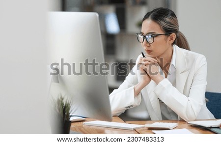 Beautiful business lady looking seriously watching on desktop monitor screen, sitting in modern office.