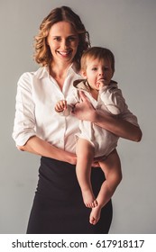 Beautiful Business Mom In Suit Is Holding Her Cute Baby Boy, Looking At Camera And Smiling, On Gray Background