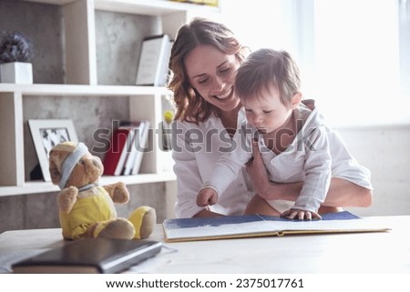 Beautiful business mom in suit is reading a book with her cute baby boy and smiling, at home