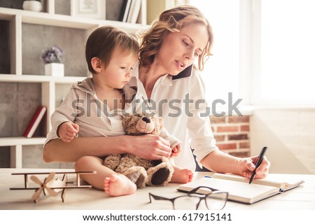 Beautiful business mom is talking on the mobile phone and taking notes while spending time with her cute baby boy at home