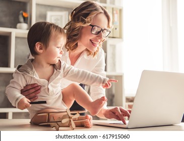 Beautiful business mom is using a laptop and smiling while spending time with her cute baby boy at home