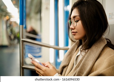 A Beautiful Business Woman Of Asian Appearance In Glasses Sits In A Subway Car And Looks At The Screen Of Her Smartphone