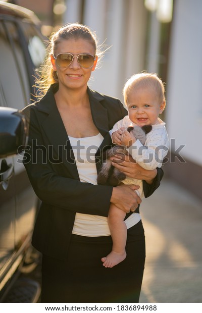Beautiful business woman mother in a suit with a\
small child in her arms against the background of her house and a\
black car goes to\
work