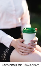 Beautiful business woman in a shirt and black skirt sits and holds a disposable paper cup with coffee in her hands close-up. Hot drink glass