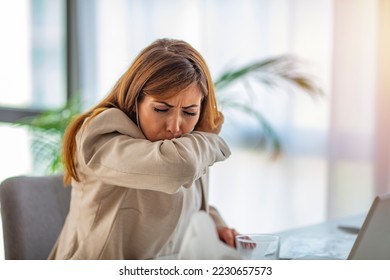 A beautiful business woman is sitting in the office, coughing in her elbow, correct behavior due to non-spreading viral infection. An unhealthy young woman feels bad at work, Covid 19 symptoms.