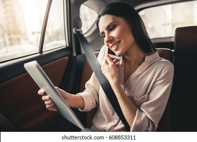 Beautiful Business Woman Is Talking On The Mobile Phone, Using A Digital Tablet And Smiling While Sitting On Back Seat In The Car