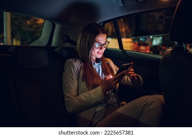 Beautiful business woman using smartphone in a car at night. Busy female working on a mobile phone in the back seat of a taxi. Bokeh and neon lights in the background. Illuminated by the screen device