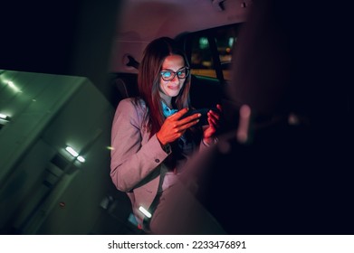 Beautiful business woman using smartphone and watching a video in a car at night. Busy female working on a mobile phone in the back seat of a taxi. Bokeh lights. View trough a windshield. Copy space.