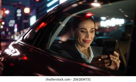 Beautiful Businesswoman is Commuting from Office in a Backseat of Her Car at Night. Entrepreneur Using Smartphone while in Transfer Taxi in Urban City Street with Working Neon Signs.