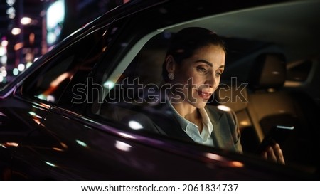 Beautiful Businesswoman is Commuting from Office in a Backseat of Her Luxury Car at Night. Entrepreneur Using Smartphone while in Transfer Taxi in Urban City Street with Working Neon Signs.