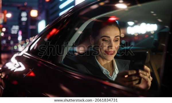 Beautiful\
Businesswoman is Commuting from Office in a Backseat of Her Car at\
Night. Entrepreneur Using Smartphone while in Transfer Taxi in\
Urban City Street with Working Neon\
Signs.