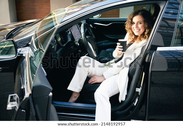 Beautiful businesswoman in elegant\
white suit sitting in modern electric car, holding cup of\
coffee.