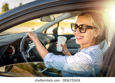Beautiful businesswoman is sitting at steering wheel of her car. She is drinking coffee and smiling. The lady is looking at the camera happily