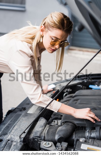 beautiful businesswoman standing\
near broken auto and looking at open trunk, car insurance\
concept