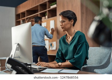 Beautiful Businesswoman Working On Desktop Computer In Her Workstation. Young Fashion Business Woman Sitting At Desk And Using Pc With Her Colleagues In Background. Woman Sitting On Workplace.