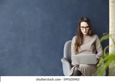 Beautiful Businesswoman Working On Laptop While Sitting In Modern Loft Office. Dark Blue Wall Background, Day Light