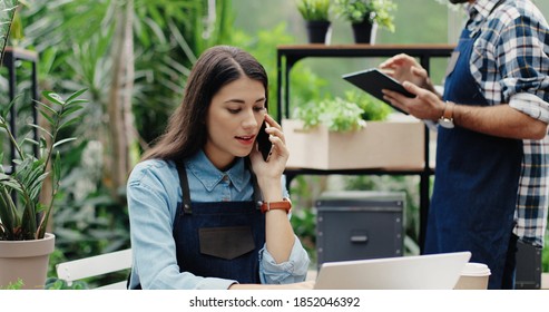 Beautiful Busy Woman Entrepreneur In Apron Sitting In Own Floral Store And Talking On Smartphone With Client. Caucasian Male Florist Typing On Tablet While Boss Speaking On Cellphone. Flower Shop