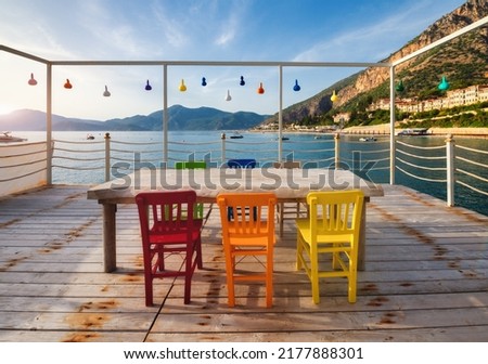 Beautiful cafe on wooden pier on the sea at sunset in summer. Landscape with colorful chairs and tables, lamps, water, mountains, beach, blue sky. Empty cozy cafe outdoor in Oludeniz, Turkey. Travel