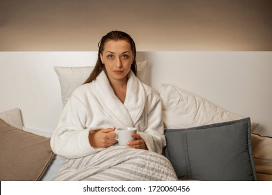 Beautiful Calm Girl With Wet Hair After Shower Relaxing At Home Sitting In Bed Drinking Herbal Tea In White Bathrobe. Young Brunette Woman Resting Before Bedtime. Beauty Treatment, Harmony Concept