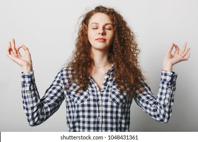 Beautiful calm young woman keeps eyes shit, stands in mudra pose, tries to relax, dressed casually, isolated over white background. Healthy female stands in lotus pose. Meditation and relax concept