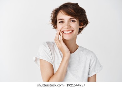 Beautiful candid woman with short hair and without makeup, touching clean facial skin and smiling, standing in t-shirt on white background.