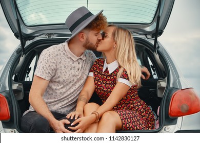 Beautiful carefree couple discovering the world traveling by car. Young man and woman kissing, hugging and touching each other while sitting in the car trunk, vehicle back view and sky in background.
