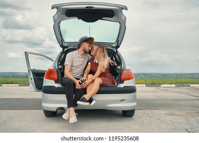 Beautiful carefree couple discovering the world traveling by car. Young man and woman kissing, hugging and touching each other while sitting in the car trunk