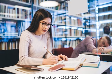 Beautiful and carefree female student in glasses reading book and taking notes while learning in bright library on blurred background 