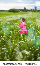 Beautiful Carefree Girl Playing Outdoors In Field With High Green Grass. Little Child Running Away From Bubbles And Laughing