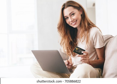 Beautiful carefree woman in casual showing credit card to camera while working with laptop on sofa