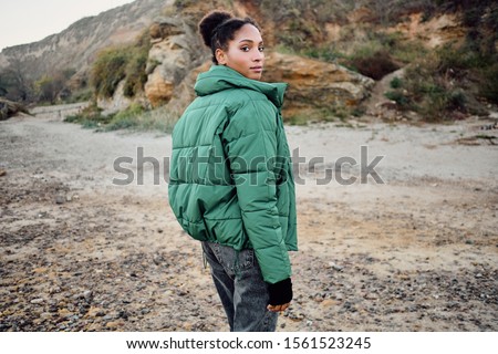 Beautiful casual African American girl in stylish down jacket dreamily looking in camera from back outdoor