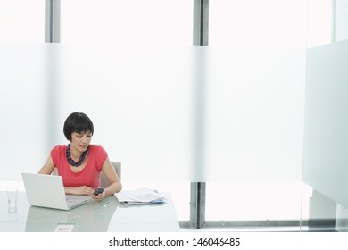 Beautiful Casual Young Woman Sitting With Laptop And Using Cellphone In Modern Cubicle At Office