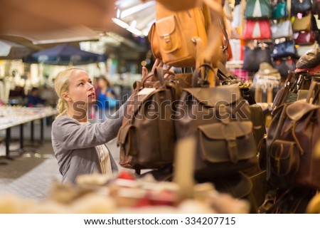 Beautiful casually dressed caucasian blond woman shopping for new leather bag on crafts market in Chania, Crete, Greece.