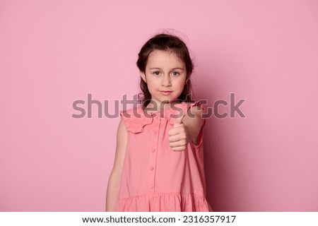 Beautiful Caucasian adorable little child girl 5-6 years old, in stylish pink dress, gestures with thumb up, expressing positive emotions, smiles looking at camera, isolated on pink background.