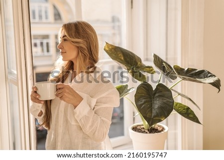 Beautiful caucasian adult woman stands at window with cup of tea or coffee in hands on light background. Blonde cheerfully starts her morning. Concept of rest and recovery.