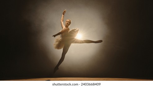 Beautiful caucasian ballerina gracefully dancing and spinning on stage, spotted by white light, isolated on smoked black background 