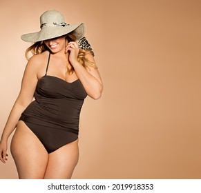 Beautiful Caucasian blond plus size woman wearing taupe swimsuit and hat in studio. Large size summer swimwear fashion.