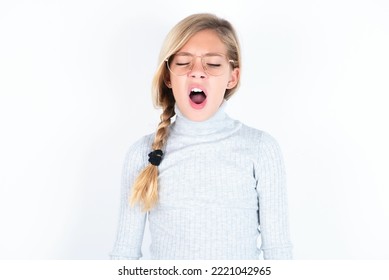 Beautiful Caucasian Blond Teen Girl Wearing Gray Turtleneck Sweater Over White Wall Yawns With Opened Mouth Stands. Daily Morning Routine