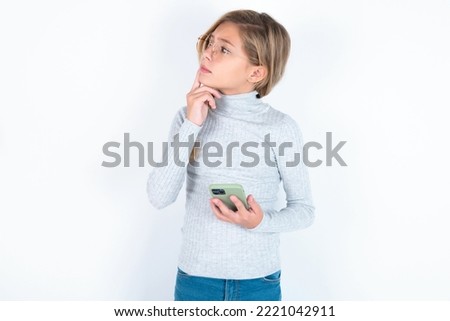 beautiful caucasian blond teen girl wearing gray turtleneck sweater over white wall thinks deeply about something, uses modern mobile phone, tries to made up good message, keeps index finger near lips