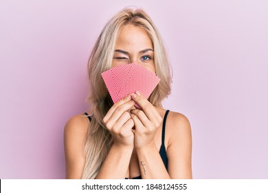 Beautiful caucasian blonde girl covering face with cards winking looking at the camera with sexy expression, cheerful and happy face. 