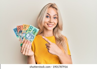 Beautiful caucasian blonde girl holding australian dollars smiling happy pointing with hand and finger 