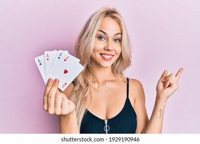 Beautiful caucasian blonde girl holding ace poker cards smiling happy pointing with hand and finger to the side 