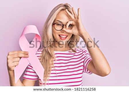 Beautiful caucasian blonde girl holding pink cancer ribbon smiling happy doing ok sign with hand on eye looking through fingers 