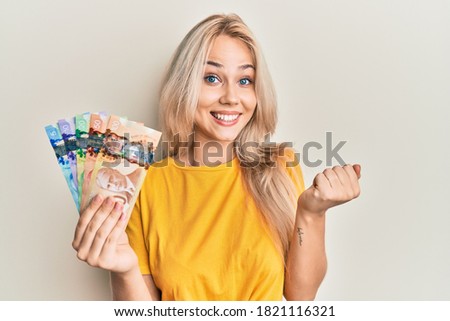 Beautiful caucasian blonde girl holding canadian dollars screaming proud, celebrating victory and success very excited with raised arm 