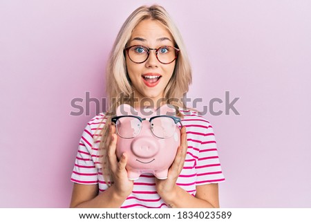 Beautiful caucasian blonde girl holding piggy bank with glasses celebrating crazy and amazed for success with open eyes screaming excited. 