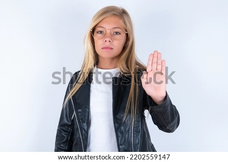 beautiful caucasian blonde little girl wearing biker jacket and glasses over white background shows stop sign prohibition symbol keeps palm forward to camera with strict expression