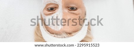 Beautiful Caucasian blonde senior woman in a modern light cosmetology center with white cream mask on a half of her face