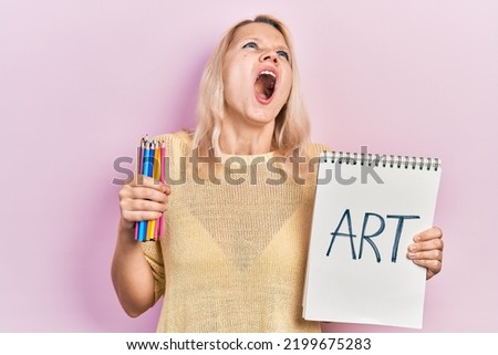 Beautiful caucasian blonde woman holding art notebook and color pencils angry and mad screaming frustrated and furious, shouting with anger looking up. 